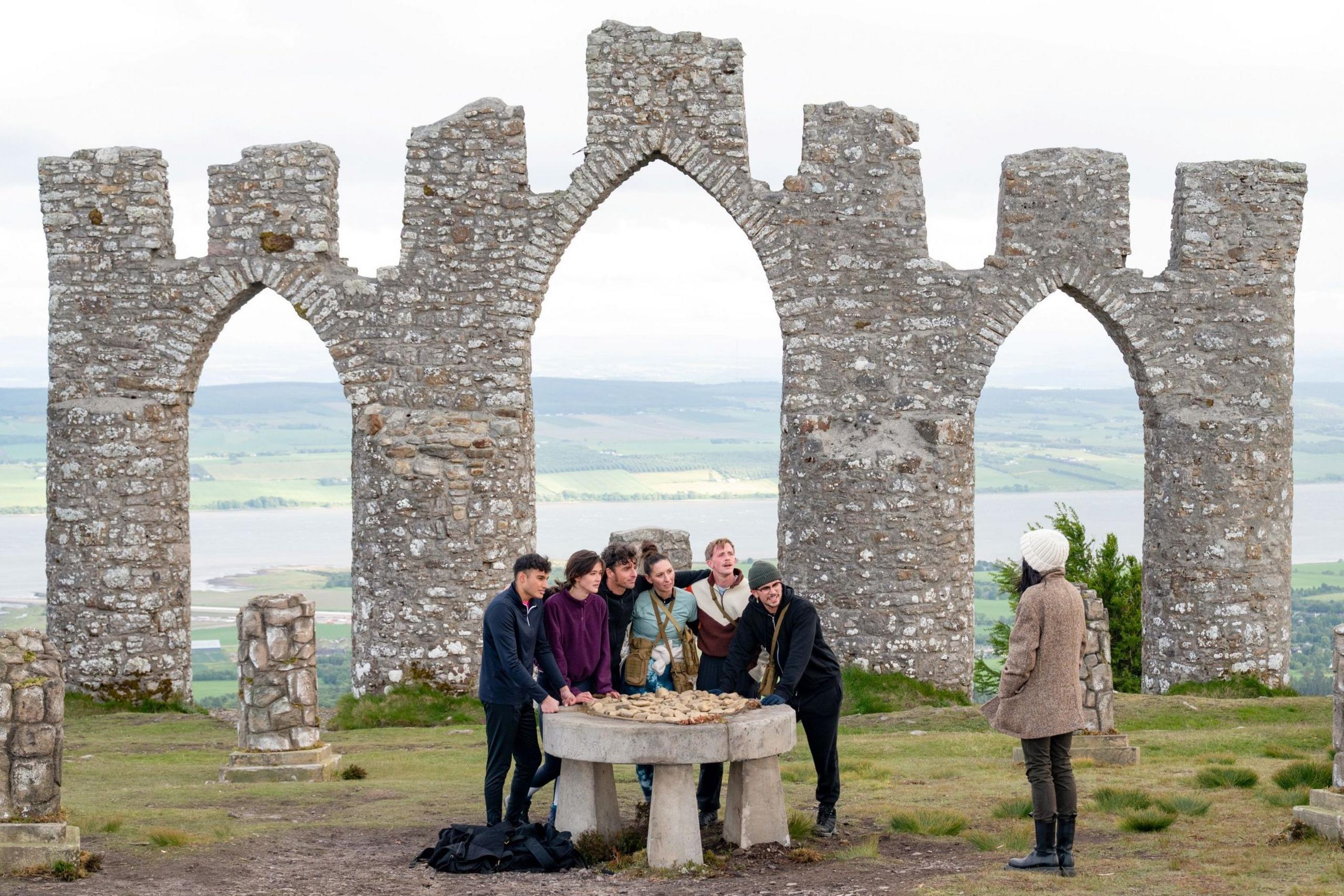 A group of contestants are gathers around a round stone table listening to Claudia Winkleman. Behind them are a series of stone arches that look like a ruin.