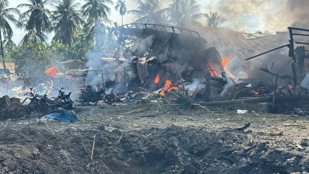 A crater in the ground is shown in the foreground as further in the distance a structure of some type is destroyed and charred, with some orange flames flickering.