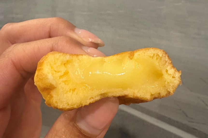 A close up of a cream puff with custard oozing. A woman with nude nails is holding the puff in her fingers.