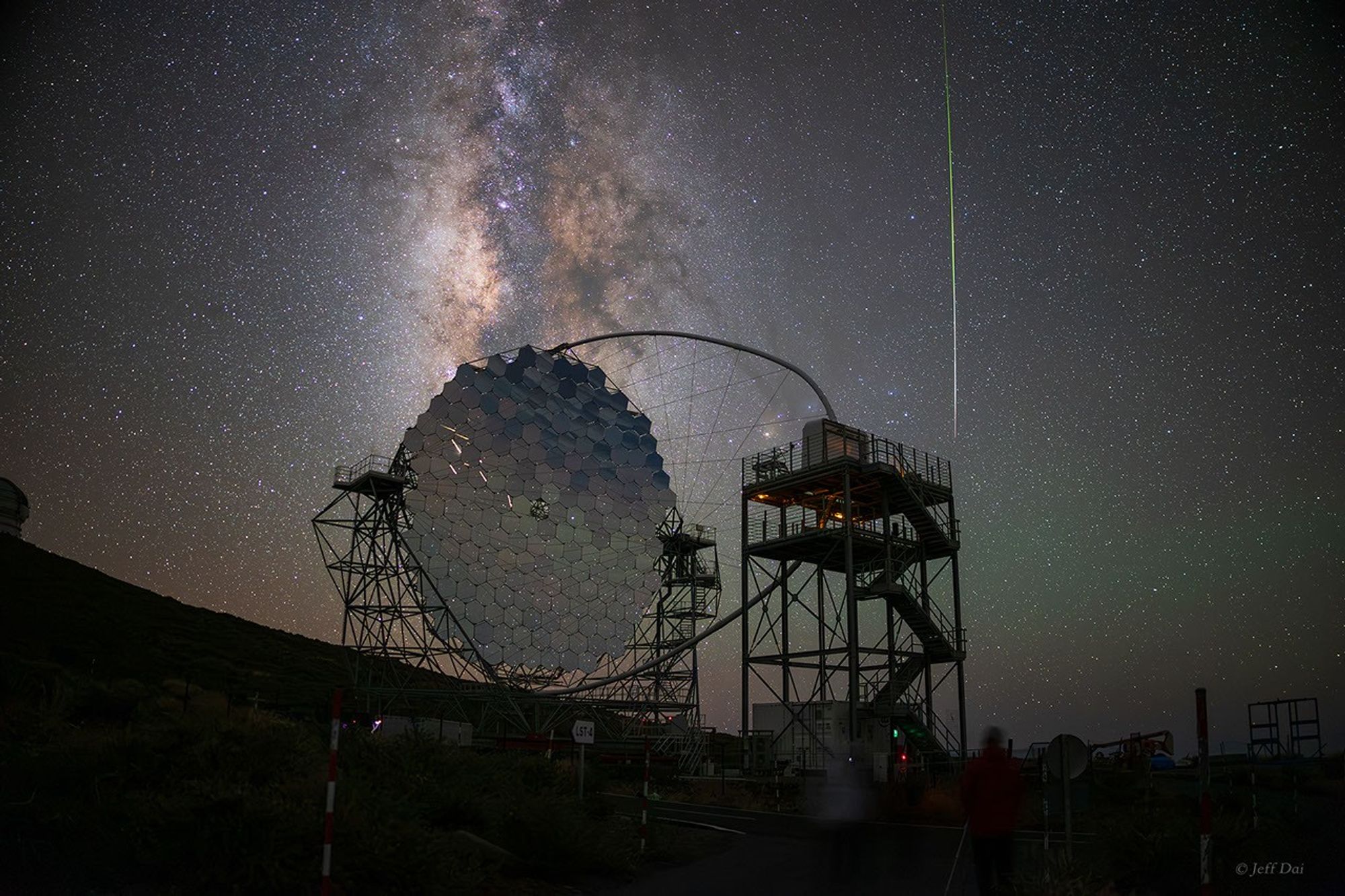 A large, segmented telescope mirror is seen against the night sky along with  the Milky Way and a bright meteor.