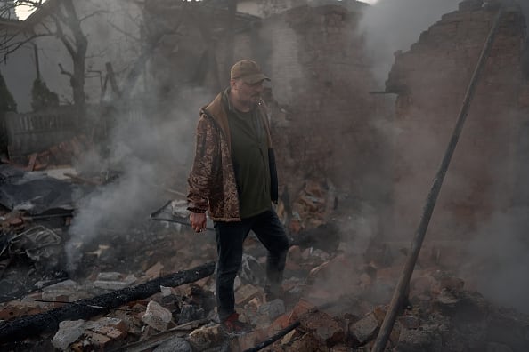 A man in a jacket and cap walks amid the burning rubble of a house at night.