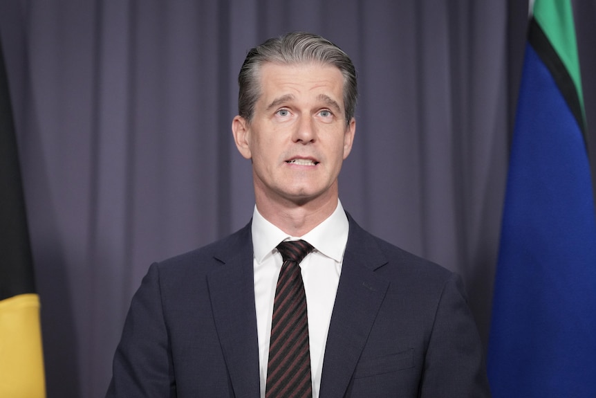 A man (Andrew Charlton) in a suit and tie stands in front of a grey backdrop with flags to the side.