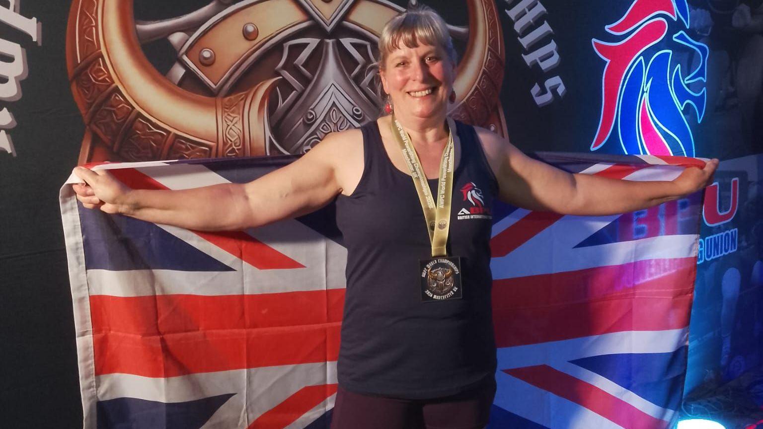 Martine Baron smiling at the camera with a medal around her neck, her arms are both stretched out wide holding a Union Jack flag. She is wearing a dark blue vest top.

