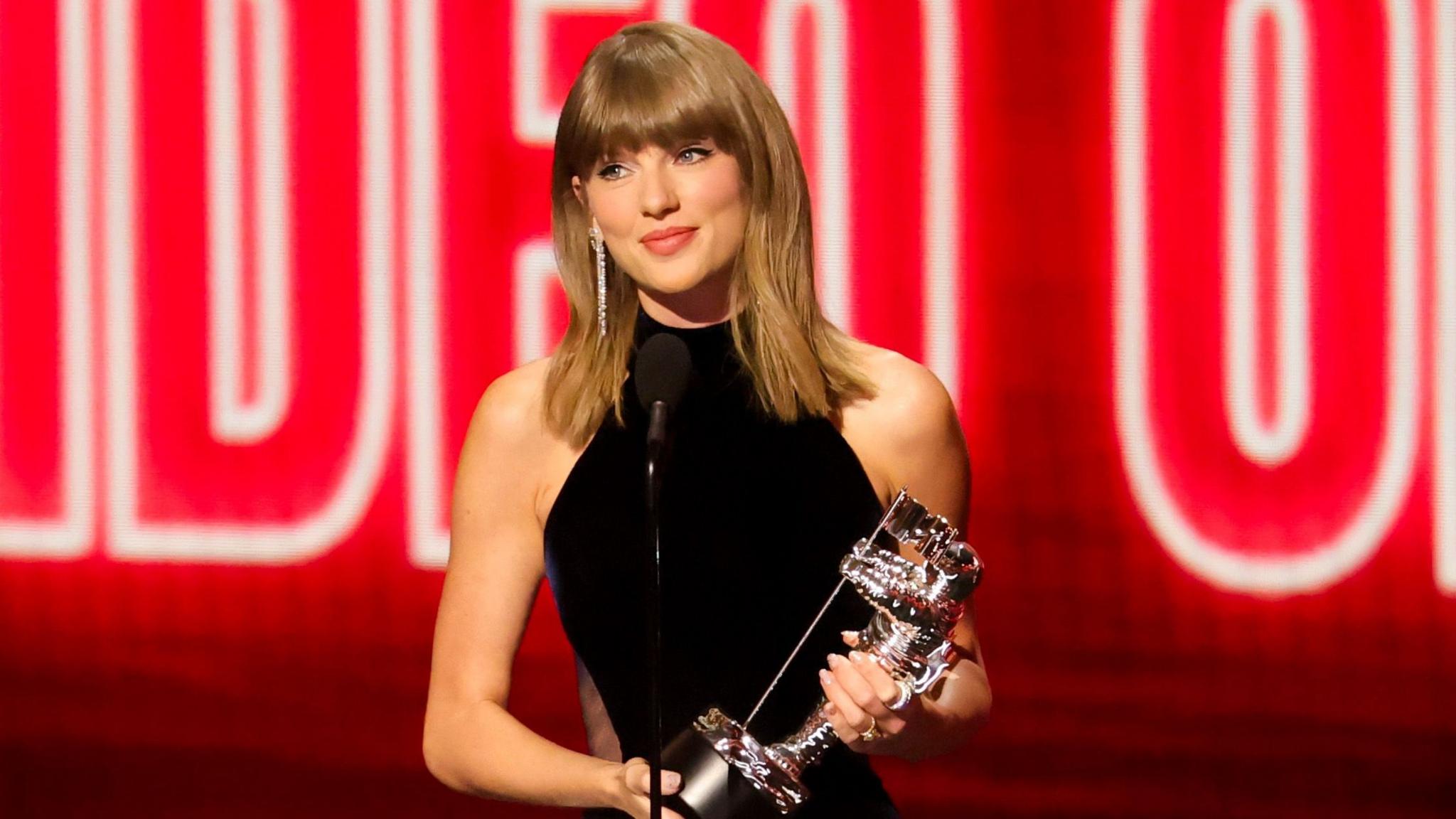 Taylor Swift is on stage accepting an award at the MTV Video Music Awards. She is holding a silver trophy and wearing a black high-neck dress. 