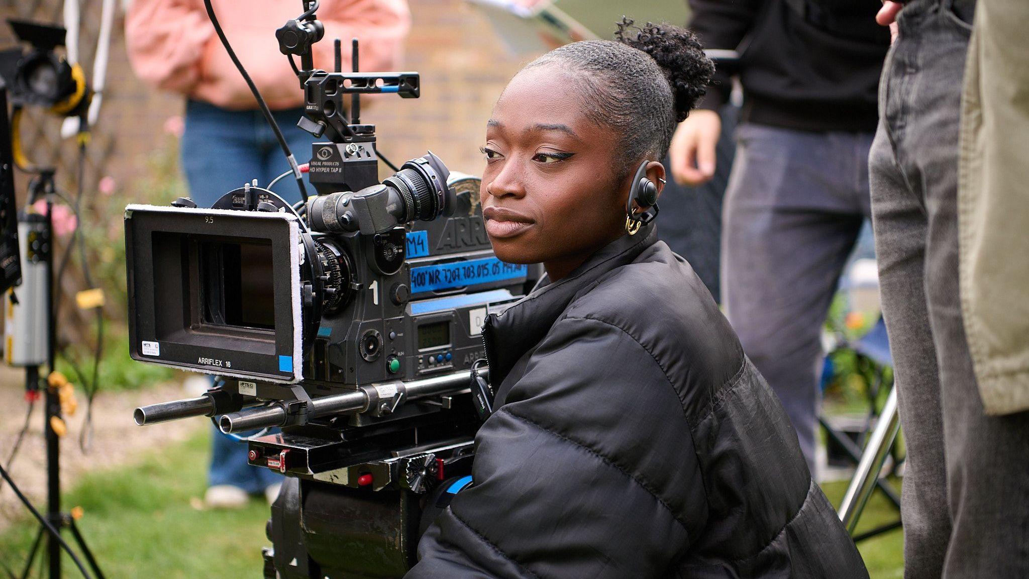 Cinematographer Melissa Cofie is holding large video camera while on set. She is wearing a black puffa jacket. 