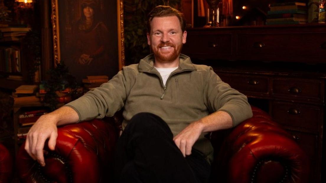 Paul Gorton is sitting in a red leather chair as he smiles at the camera ahead of a podcast recording for The Traitors. 