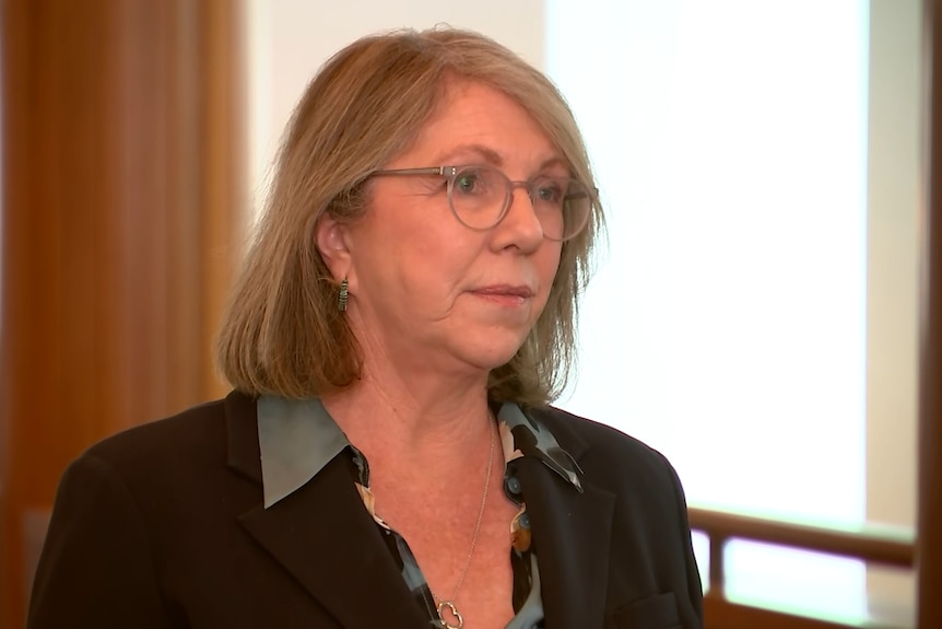 Ms King in a black blazer and glasses looking serious at reporters at a press conference.