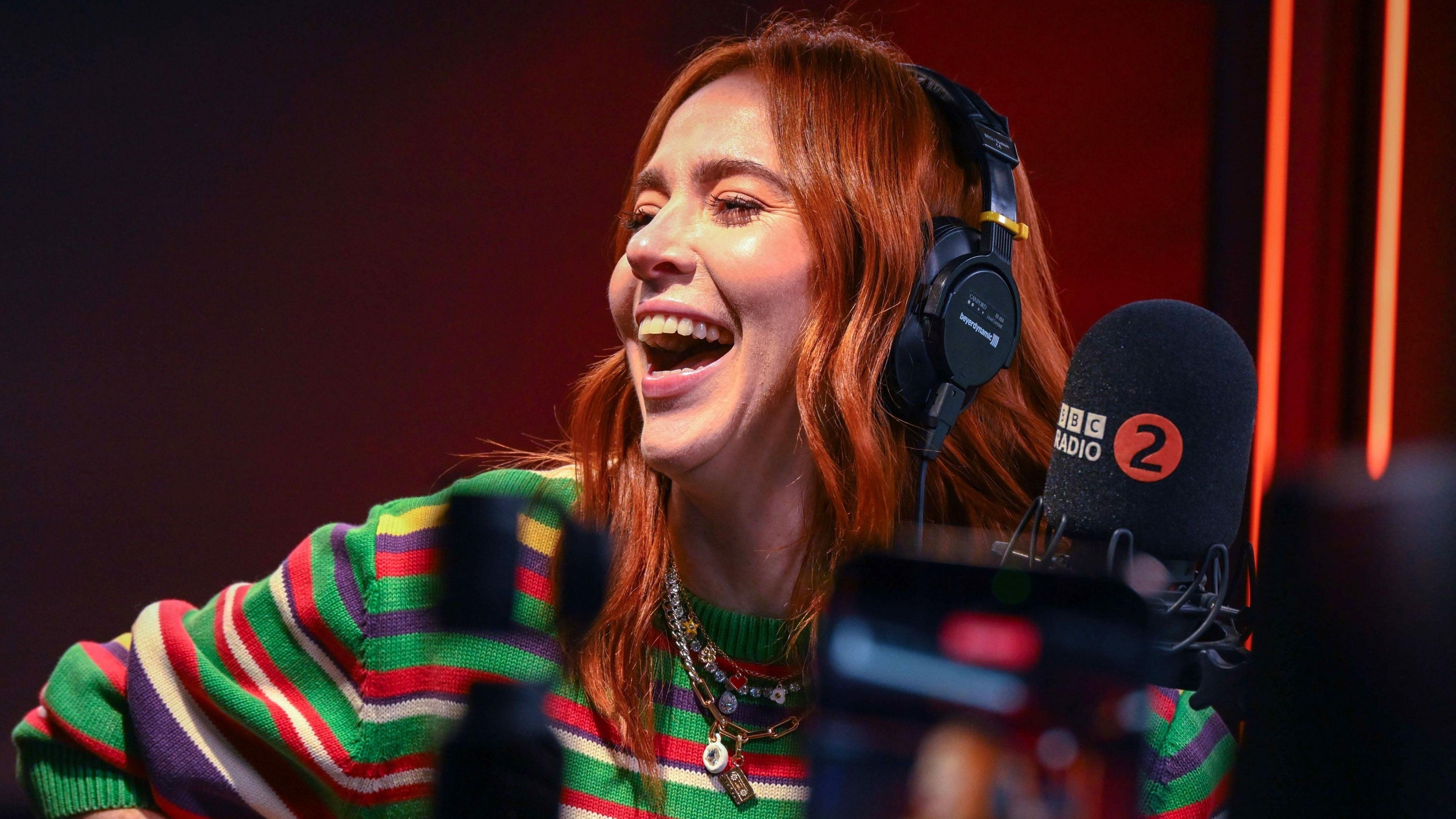 Angela Scanlon laughing, wearing headphones in a radio studio front of a microphone with the BBC Radio 2 logo